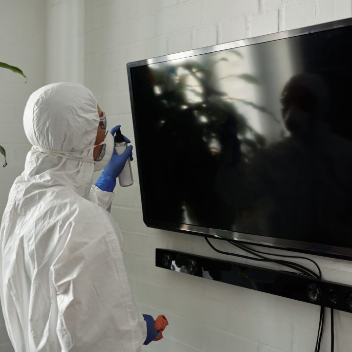 A person wearing PPE disinfecting a TV screen inside a room during the COVID-19 pandemic.